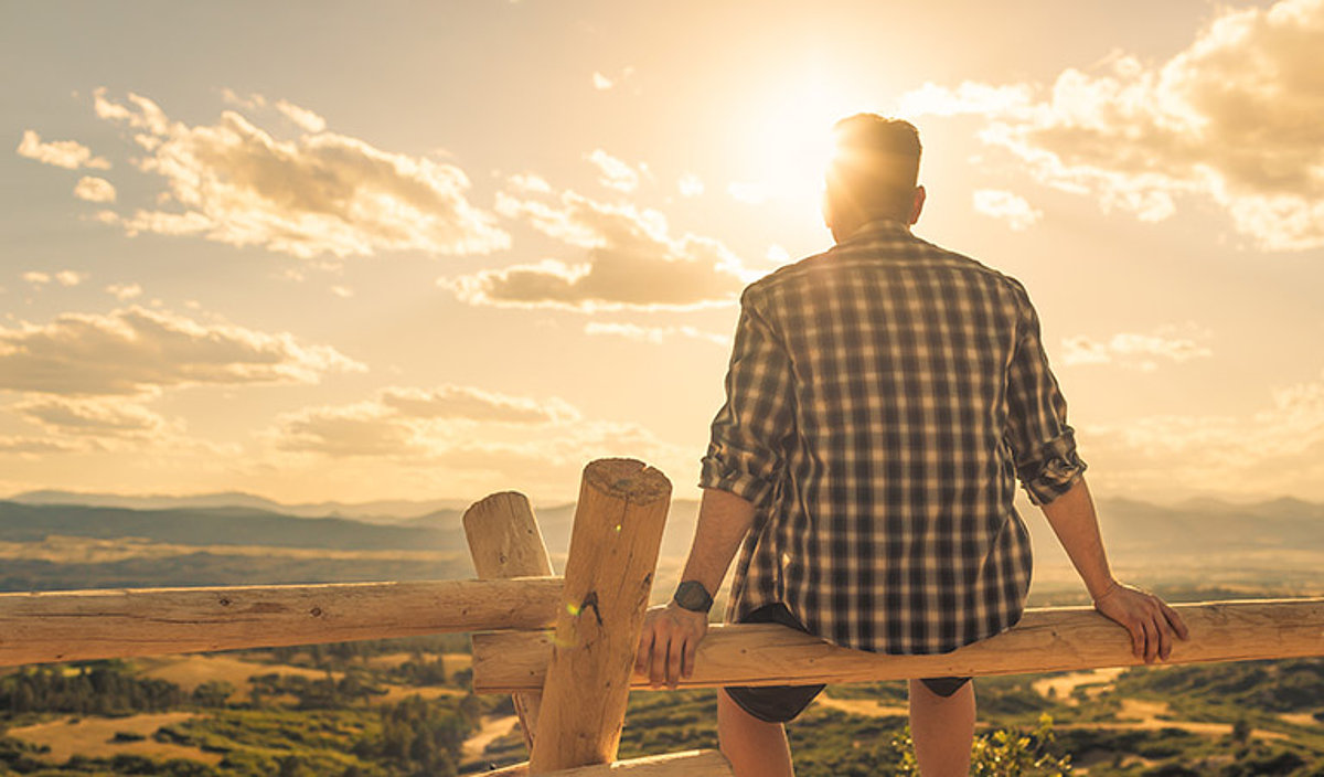 Eine männliche Person sitzt auf einem Holzzaun und blickt gen Sonne