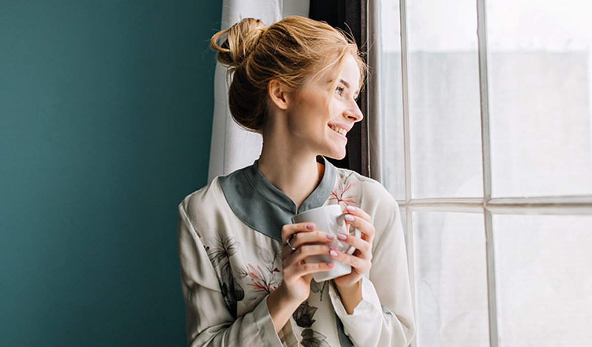 Junge Frau blickt aus dem Fenster und hält eine Tasse Tee in beiden Händen